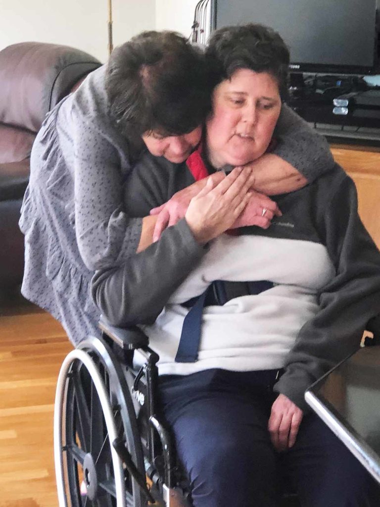 Kim sits in wheelchair while getting hugged from behind by a loved one.