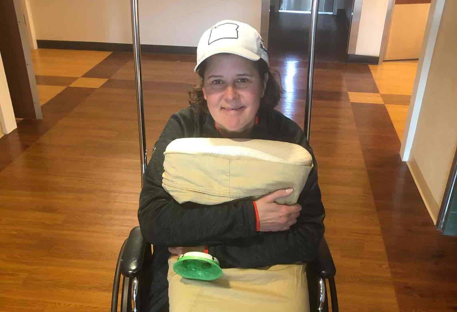 Kim sits in a wheelchair in a hospital hallways while hugging a pillow and smiling for the camera.